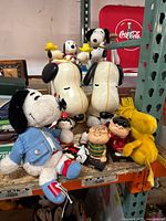Wide angle of plush and plastic Peanuts character toys including large Snoopy figures and small figurines on a metal shelf