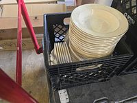 Photo showing a stack of large white deep dish bowls with smaller matching bowls inside a black crate, boxes and miscellaneous items around.