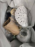 Box containing various home decor items including glassware, white crochet doily, figurine, and wrapped vase.