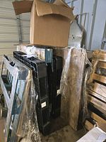 Photo showing stacked and wrapped black plastic pallets or parts along with several used wooden pallets and cardboard boxes on top in a warehouse setting.