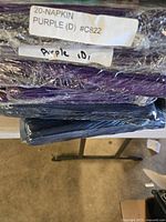 Close-up showing stacked wrapped purple napkins labeled 20-NAPKIN PURPLE (D) #C822 and Purple ID1 in plastic wrapping, sitting on table surface.