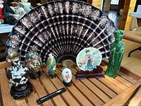 Photo showing nine Asian-inspired decorative items including fan, porcelain figurines, cloisonné vase, brass vase, and chopsticks.