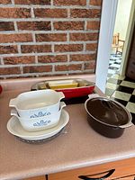 View showing multiple baking dishes including Pyrex glass dishes with blue floral patterns, beige CorningWare ceramics, and Le Creuset stoneware pieces in brown and red.