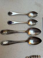 Four sterling silver spoons arranged side by side, showing two smaller spoons with enamel crests and two larger spoons with lined handles.