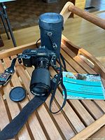 Canon T70 film camera with zoom lens, lens cap, woven camera strap, cylindrical carrying case, and manuals on wooden surface.