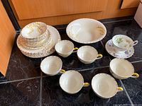 Full set of bone china plates, cups, saucers, and serving bowl arranged on surface. Most pieces show yellow/brown leaf motif, one cup and saucer have floral design.