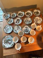 Entire 16-piece ceramic dinnerware set displayed on wooden table under natural light. Items include bowls, plates, cups, and saucers featuring The Friendly Village pattern from Johnson Bros.