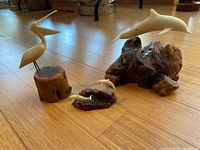 Set of three John Perry animal sculptures on wooden floor: one bird sculpture mounted on a wooden log base and two dolphin sculptures (one single and one duo) mounted on darker wood bases.