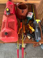 Photo shows a collection of garage and workshop tools and accessories on a wooden table including a red plastic gas cans, cordless power tool, screwdrivers, clamps, and other small items