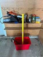Overview photo of power tools and hardware on pegboard and shelf, with red bucket holding sledgehammer and crowbar on floor