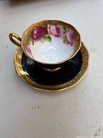 Top view of teacup showing detailed pink rose floral pattern with gold rim and gold handle, sitting on matching black and gold saucer