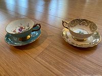 Side angle of both teacup and saucer sets showing turquoise and gold floral design on the Paragon set and golden filigree on the Hammersley set.