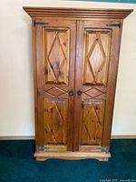 Front view of solid wood pine armoire showing double doors with diamond and cross pattern panels, metal hinges, ring-pull handles, and layered crown molding on top.