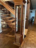 Front angled view of the tall wooden and glass curio cabinet placed near a wooden staircase on a parquet floor.