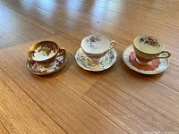 Three teacup and saucer sets arranged in a row on a wooden floor, showcasing varied floral designs and gold accents.