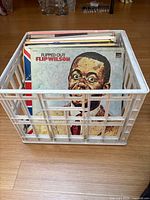 White plastic milk crate containing multiple vinyl records with Flip Wilson 'Flipped Out' album prominently visible at front.