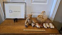 Photo showing a wooden table with a large textured light brown teddy bear, five smaller teddy bears in a row, and a woven basket to the right. A ruler placed in front shows scale.