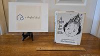 Front view of 'A Light in the Attic' book with dust jacket, placed next to a ruler to show size, alongside a logo sign.