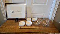 Full set of four white ceramic ramekin bowls and four clear glass bowls on wooden table with a framed sign behind them.