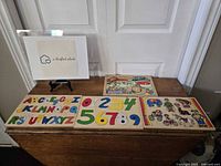 Four vintage wooden puzzles displayed on a rustic chest, including alphabet, numbers, Fisher Price animal, and motorcycle rider puzzles