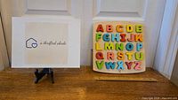 Photo showing the complete Hape Wooden Alphabet Puzzle on wooden surface next to a sign with logo.