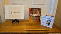 Two books displayed on a wooden surface: "An Introduction to Painting Portraits" and "Playing with Art", with a small framed sign.
