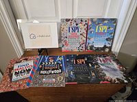 Photo showing all seven books in the lot displayed on a wooden chest with a business card on a stand. Titles include four I Spy books and three Where's Waldo puzzle books.