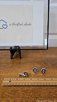 Photo showing the cufflinks and tie clip arranged on a wooden surface with a ruler for size reference and a display sign in the background.