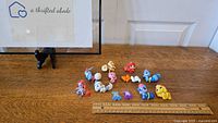 Nine colorful Littlest Pet Shop animal toy figures and two additional small characters arranged on wooden table with ruler for size reference.