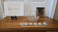 Full frontal photo showing the six Itty Bitty Critter pig figurines aligned in front of a ruler, a clear cut crystal bowl centered behind them, and a rectangular carved wall plaque to the right, all placed on a wooden surface.