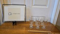 Six Glastonbury Lotus clear stemmed footed parfait glasses arranged on a wooden surface next to a framed sign and a wooden ruler for scale.