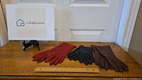 Three pairs of vintage leather ladies gloves in red, black, and brown beside a ruler showing length, on a wooden surface.