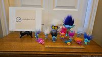 12 assorted Trolls movie character toys arranged on a wooden surface in front of a white door. The toys have colorful synthetic hair and detailed features.