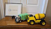 Two Tonka toy trucks on wooden surface - a green recycling garbage truck and a yellow cement mixer truck.