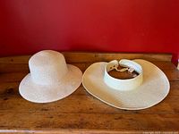 Both summer hats displayed on a wooden surface against a red wall. Left hat is pale pink cloche style, right hat is beige open-top visor with wide brim.