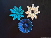 Three vintage enamel flower brooches displayed on black background: teal double-petal, white and black daisy, and royal blue daisy.