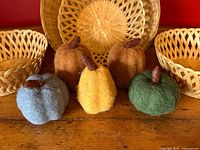 Four woven baskets in natural finish displayed with five colorful felted wool gourds in green, yellow, brown, rust, and blue colors with wool stems on a wooden surface.