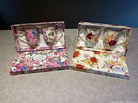 Photo showing two boxed sets of fine china mugs from ACE Gift Collection. Left box contains mugs with pink and blue rose designs, right box contains mugs with red, yellow, peach rose designs. Satin lining in boxes visible.