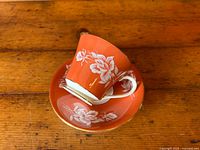 Photo showing the coral red teacup stacked inside the matching saucer with white floral decoration and gold trim on both pieces.