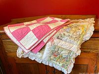 Two handmade baby quilts folded and draped over a wooden cabinet, showing the pink and white checkerboard pattern and the cream quilt with colorful animal illustrations and lace trim.