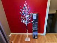 Full view of the 5-foot white artificial tree with colorful LED lights lit, along with the original box standing beside it against a red wall.