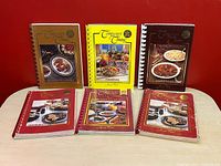 Top-down view of six spiral-bound cookbooks by Jean Paré from the "Company's Coming" series arranged on a table against a red background.