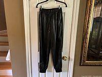 Full length photo of black faux leather jogger pants hanging on black hanger, showing elastic cuffs at the ankles.
