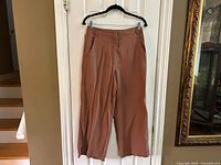Full view of rust brown wide-leg trousers hanging on black hanger against white door. Shows length, cut, and color of the garment.