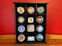 Black wooden display cabinet holding 12 miniature porcelain plates, each with distinct floral and ornamental designs inspired by imperial court artistry.