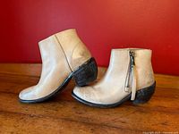 Side profile showing beige leather upper, almond toe, stacked heel, and side zipper on boots placed on wooden floor against red wall.