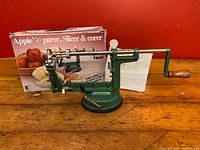 Apple peeler, corer & slicer unit with wooden handle on suction base, displayed on wooden surface with original box and instruction sheet behind it.