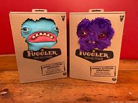 Front view of two sealed Fuggler Funny Ugly Monster plush toys in their original die-cut cardboard boxes, showing the monsters' faces through the cutouts.