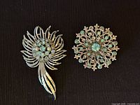 Pair of vintage mid-century brooches on dark background, showing detailed metalwork and rhinestones with aqua and pale blue tones.