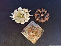 Top view showing three brooches against a black background: enamel flower with rhinestone center in white and pale green, copper-tone rounded flower, and square lucite mosaic brooch.